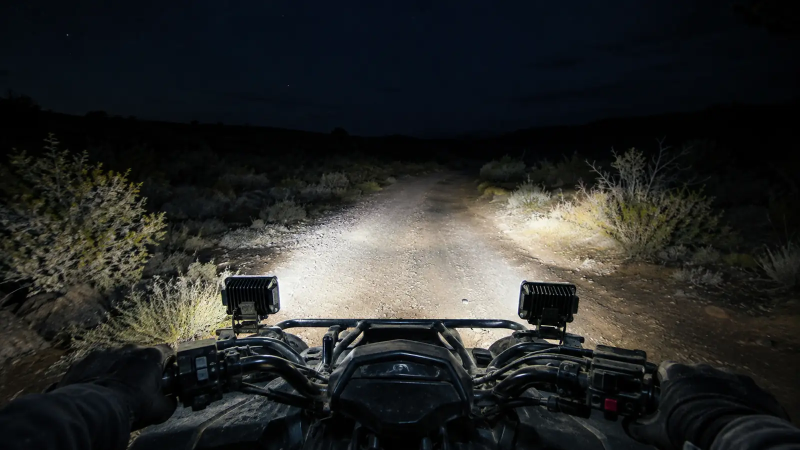 An ATV on a dark trail at night with two LED pods lit and the beam reaching down a dirt road